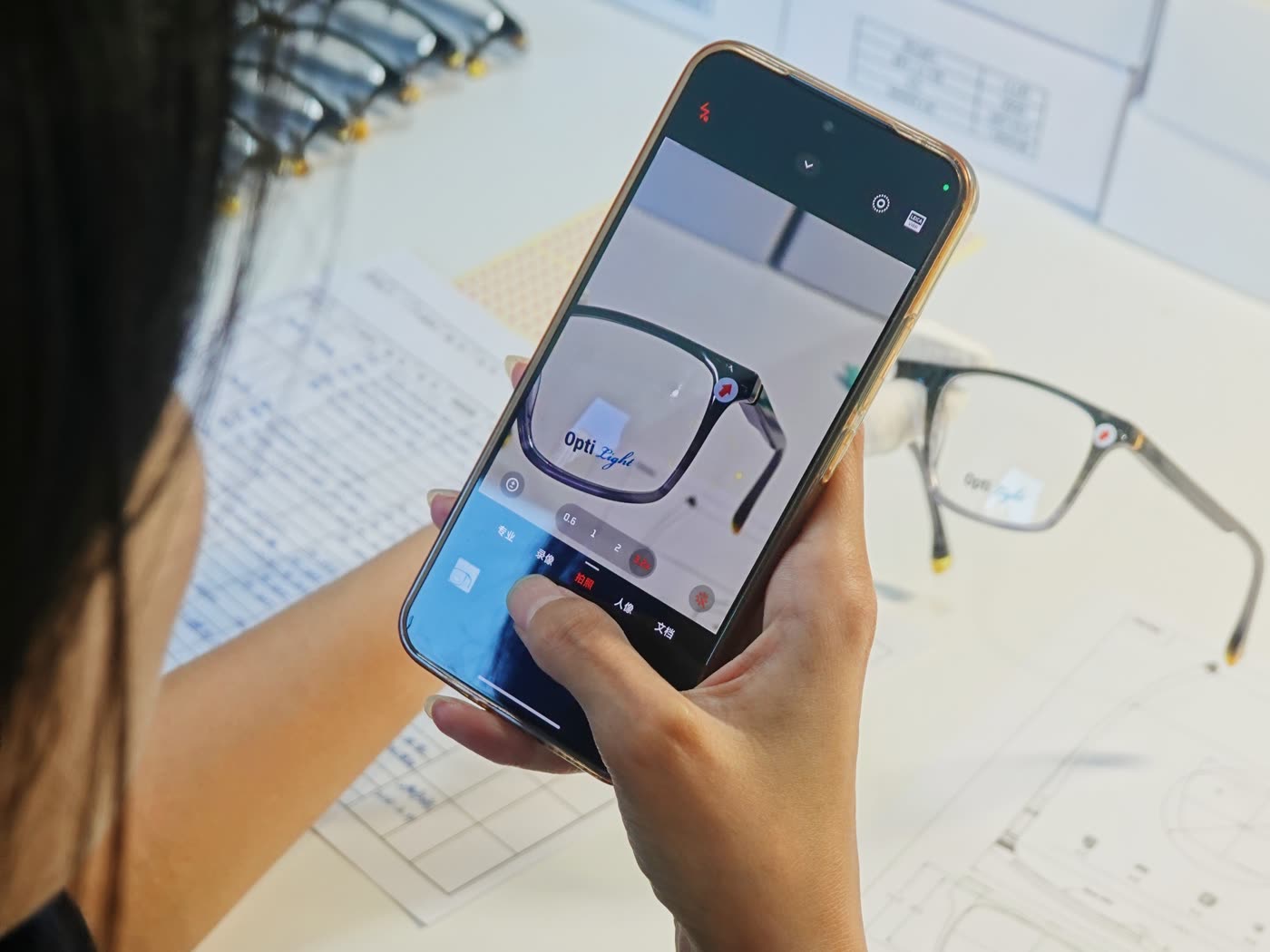 A gloved hand holds a frame while the other hand photographs it with a phone, over inspection sheets and a technical drawing