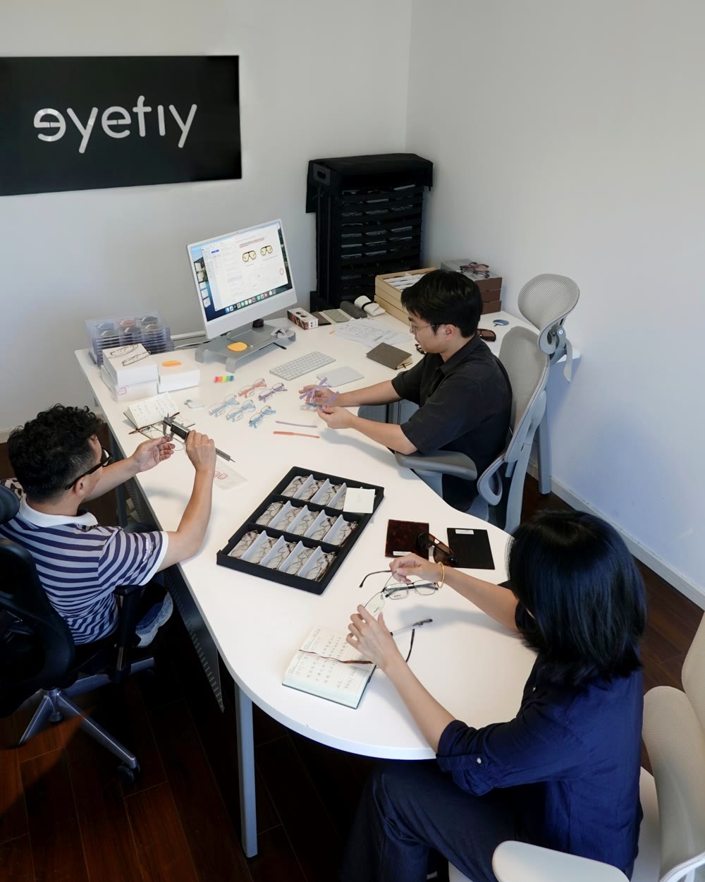 Three people at a shared table reviewing eyewear samples: a tray of frames, a vernier caliper, drawings on screen