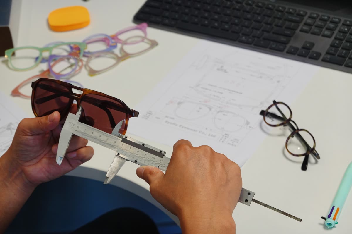 Hands measuring a sunglass frame with a vernier caliper over the technical drawing it is checked against
